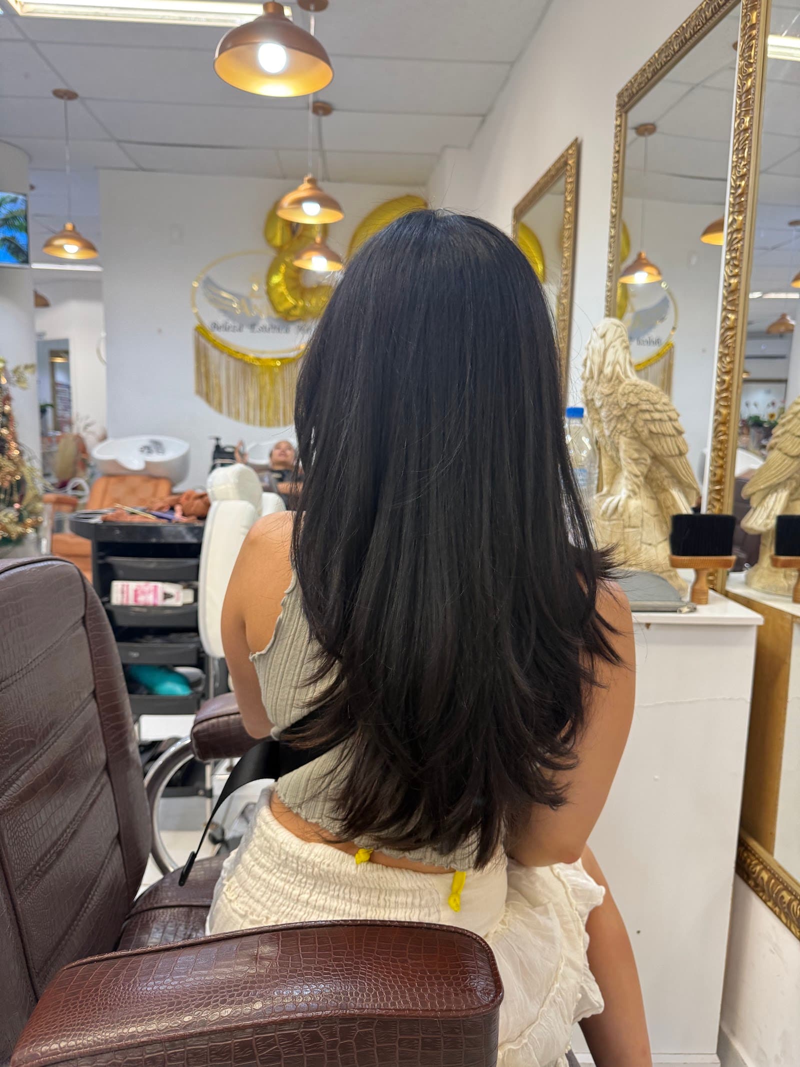 Cliente de cabelo preto longo e liso, de costas, no salão do Zu Anjos Hair, entre espelhos de moldura dourada e pendentes acesos