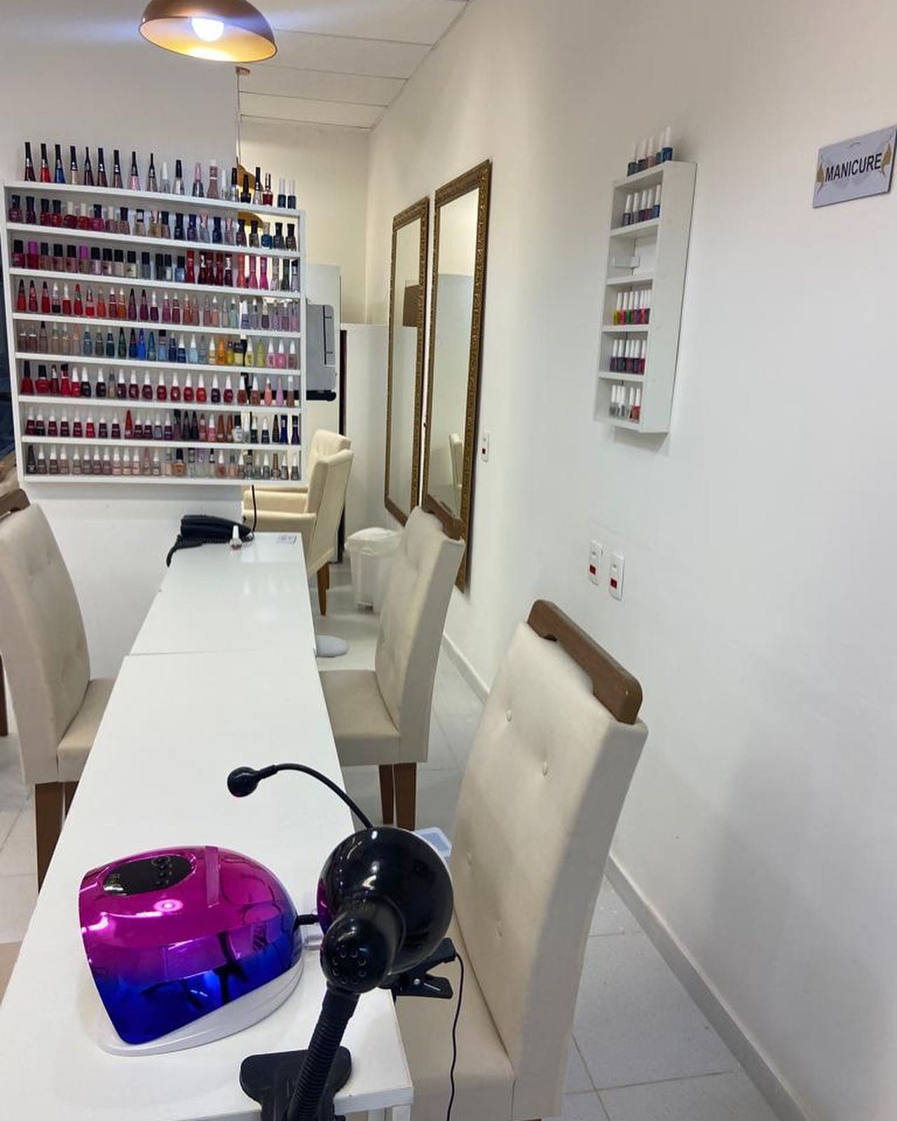 Bancada da manicure com cadeiras claras e a parede de esmaltes ao fundo