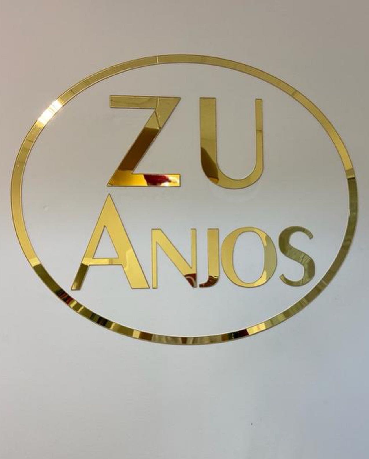 Logo do Zu Anjos em letras douradas espelhadas dentro de um oval dourado, na parede branca do salão